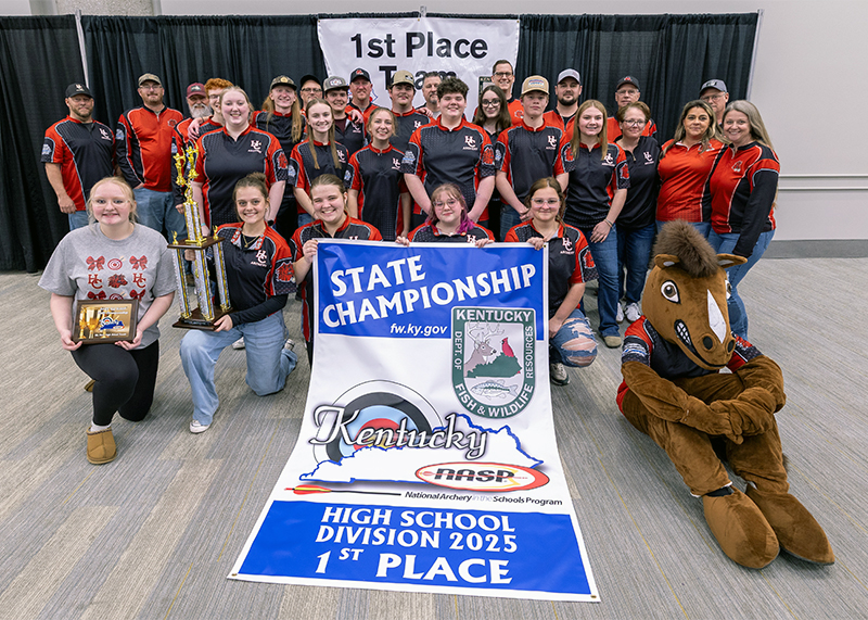 A student is holding a genesis bow and trophy and standing in front of a sign that reads Individual Awards