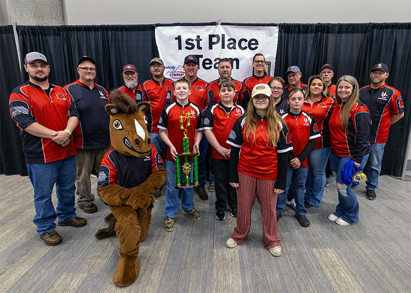 A group of four students and thirteen adults in front of a sign that reads 1st place team
