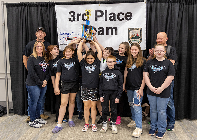 a group of students and instructors are standing for a group photograph. In the center, a student is holding a large trophy above her head. The sign behind them reads 3rd place team