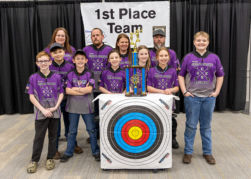 A group of students are standing in front of a sign that reads 1st place team. the student in the center is holding up a large trophy and two students are holding up a archery target