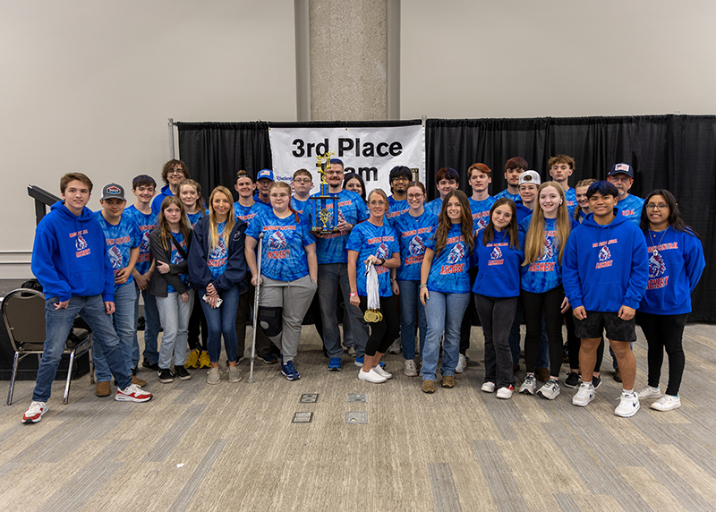 a group of students and instructors are standing for a group photograph. In the center, a student is holding a large trophy above her head. The sign behind them reads 1st place team