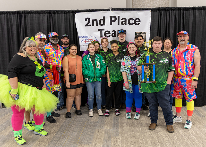 A group of students are standing in front of a sign that reads 2nd place team. the student in the center is holding up a large trophy