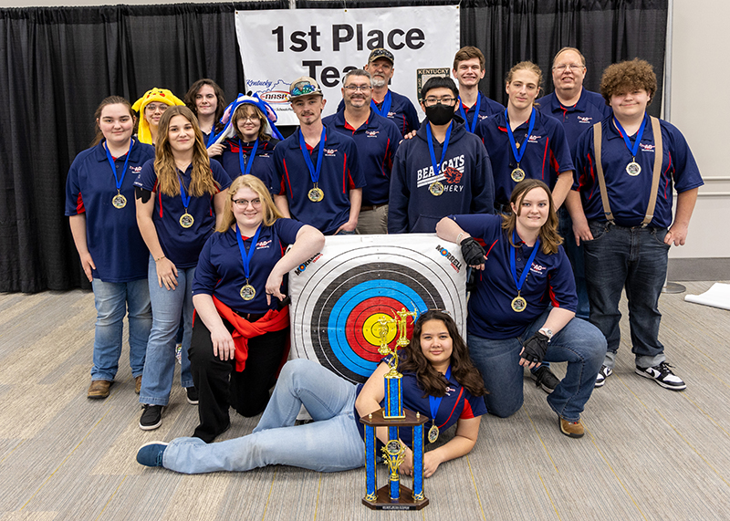 A group of students are standing in front of a sign that reads 1st place team. the student in the center is holding up a large trophy and two students are holding up a archery target