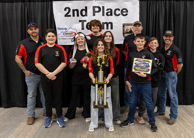 a group of students and instructors are standing for a group photograph. In the center, a student is holding a large trophy above her head. The sign behind them reads 2nd place team