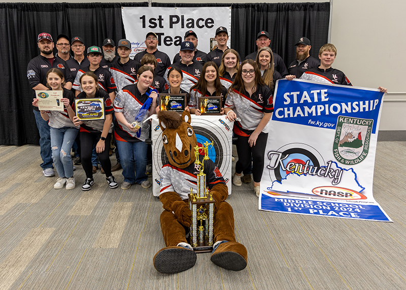 a group of students and instructors are standing for a group photograph. In the center, a student is holding a large trophy above her head. The sign behind them reads 1st place team