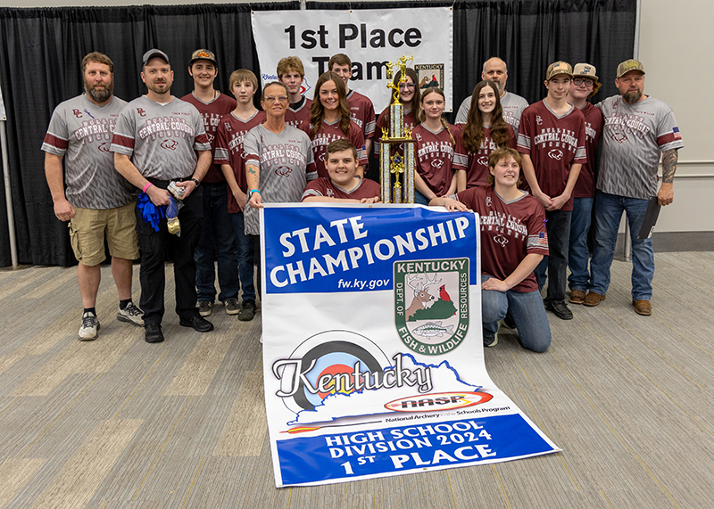 A group of students are standing in front of a sign that reads 1st place team. the student in the center is holding up a large trophy and two students are holding up a archery target