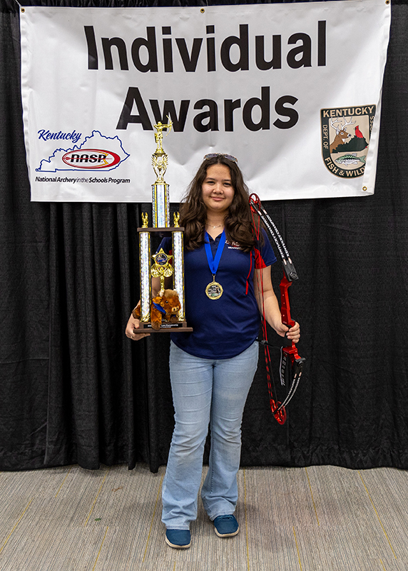 A student is holding up a large trophy and genesis bow. Behind her a sign reads individual awards