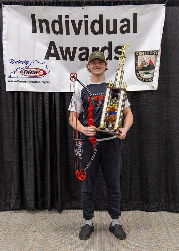 A student is holding a genesis bow and trophy and standing in front of a sign that reads Individual Awards