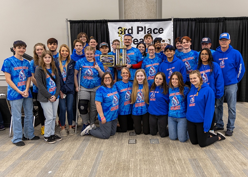 a group of students and instructors are standing for a group photograph. In the center, a student is holding a large trophy above her head. The sign behind them reads 3rd place team