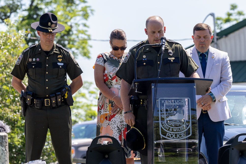 An officer is speaking at a podium as another officer, a woman and the commissioner stand behind him with their heads down in prayer