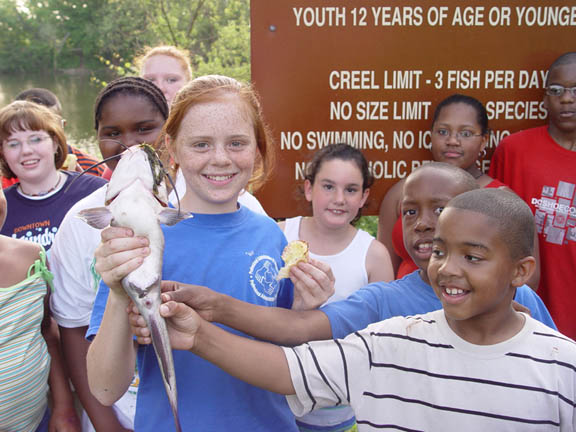 Kids standing in front of a fishing regulations sign holding a catfish and smiling. Background sign includes age, creel, and size limits. Boater Resources