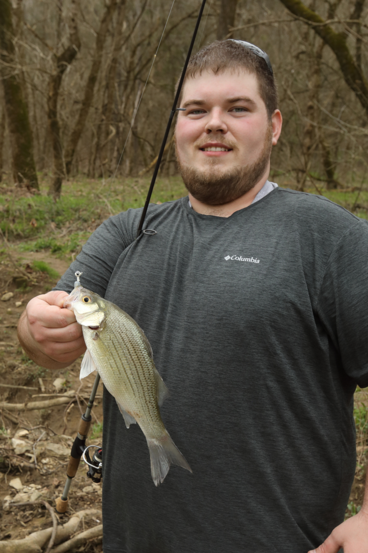 A Young man is holding up a white bass in his left hand and a fishing pole in his right