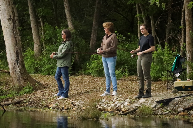 Three women are standing on an edge of a pond and fishing