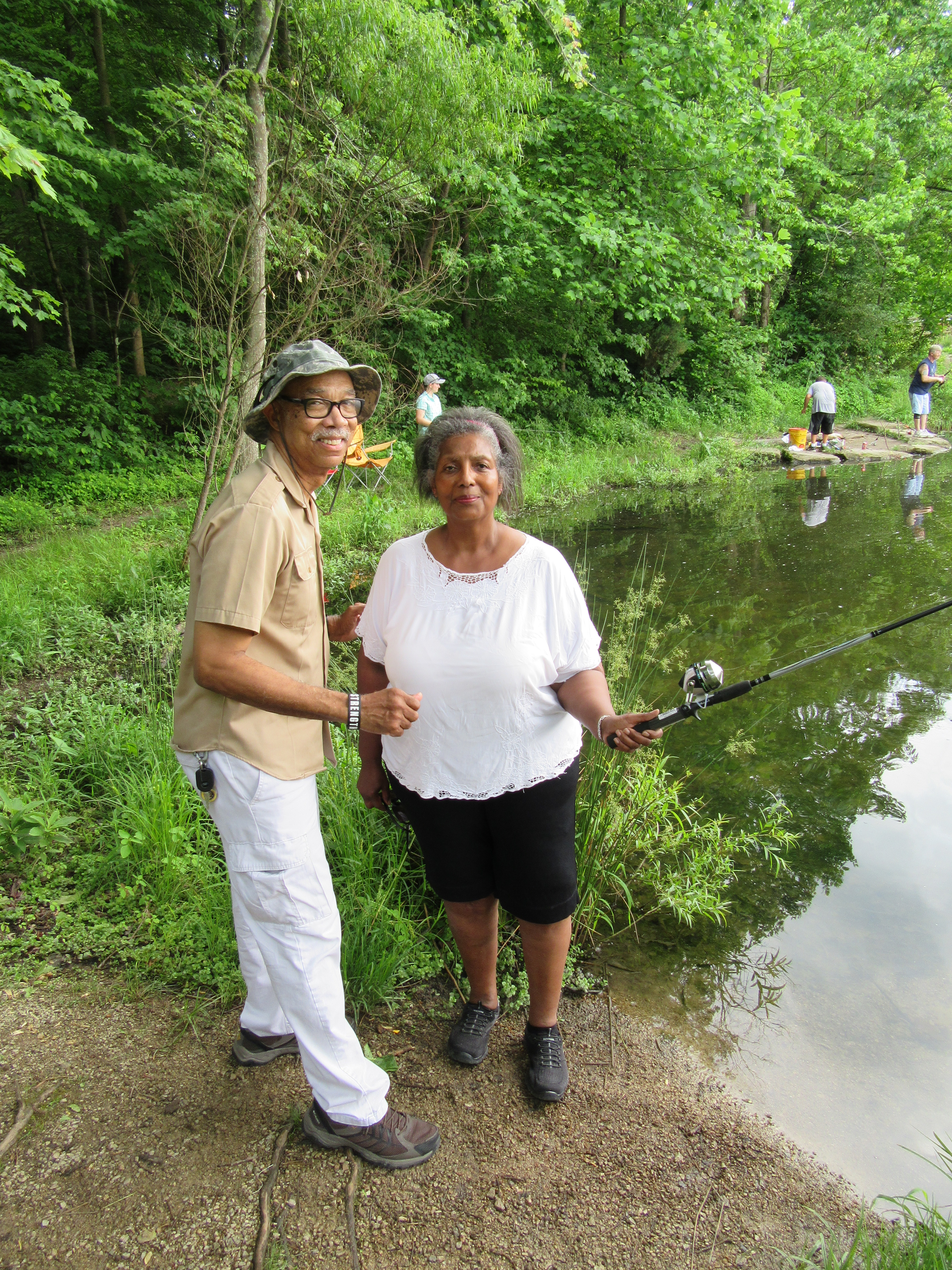 A couple is smiling for the camera as a woman is holding out a Fishing Pole into a pond